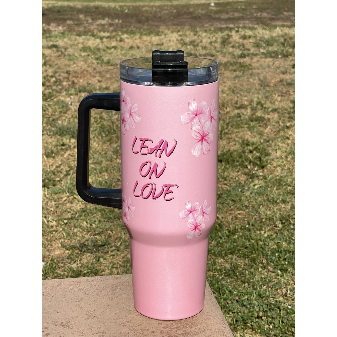 Alpha Kappa Alpha Ivy Love Sisterhood Tumbler with Handle (Lifestyle) - A pink insulated tumbler with a black handle and lid, featuring the text 'LEAN ON LOVE' and pink cherry blossom illustrations, set outdoors on a concrete surface with grass in the background.