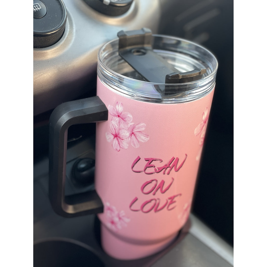 Alpha Kappa Alpha Ivy Love Sisterhood Tumbler with Handle (Lifestyle) - A pink insulated travel tumbler with a black handle and clear lid, featuring cherry blossom graphics and the text 'LEAN ON LOVE', shown resting in a car cup holder.