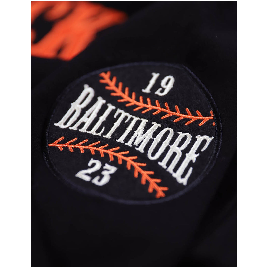 Baltimore Black Sox Heritage Hooded Sweatshirt (Sleeve Detail) - Close-up of a black baseball patch on a dark garment, featuring white 'BALTIMORE' lettering, orange stitched seam details, and the numbers 19 and 23.