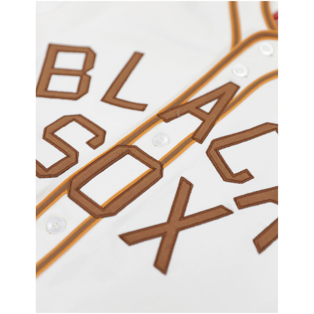 Baltimore Black Sox Negro Leagues Heritage Baseball Jersey (Front Detail) - Close-up of a white baseball-style top with large brown embroidered letters spelling part of a word, outlined with gold piping and white buttons.