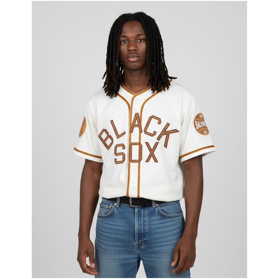 Baltimore Black Sox Negro Leagues Heritage Baseball Jersey (Lifestyle) - A man modeling a vintage-style off-white baseball jersey with 'BLACK SOX' in brown lettering outlined in gold, paired with blue jeans, against a solid gray background.