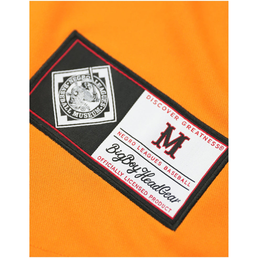 Baltimore Black Sox Legacy Baseball Jersey (Front Detail 3) - Close-up of an orange athletic jersey with a rectangular embroidered patch. The patch is black and white with red trim, featuring a Negro Leagues Baseball logo on the left and a large red 'M' with 'BigBoyHeadGear' and 'DISCOVER GREATNESS' text on the right.