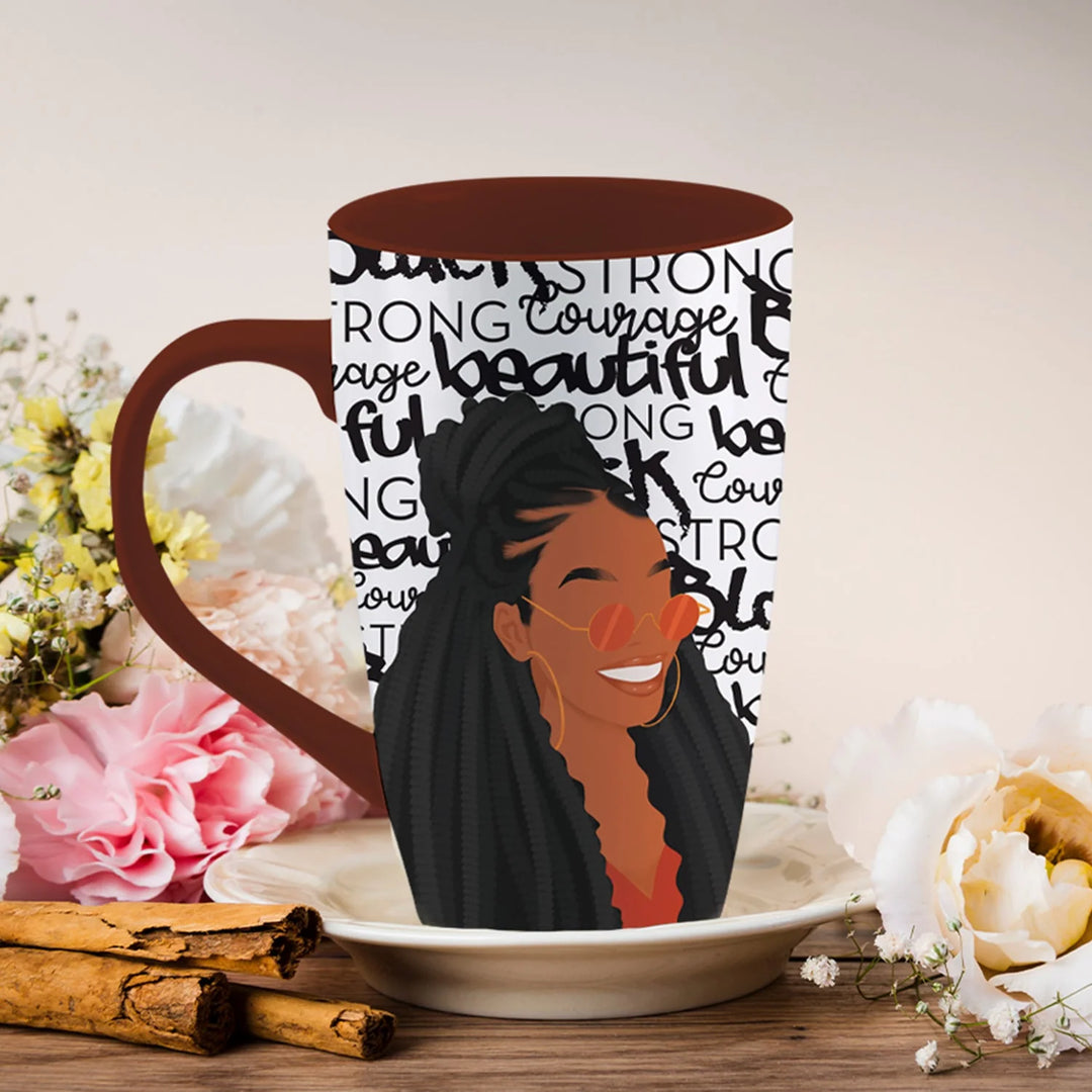 Black and Beautiful Ceramic Latte Mug (Lifestyle) - A ceramic coffee mug with a brown handle and rim, featuring a black and white typographic background with an illustration of a smiling Black woman with glasses. The mug sits on a white saucer on a wooden table, surrounded by pink and white flowers, cinnamon sticks, and a neutral background.