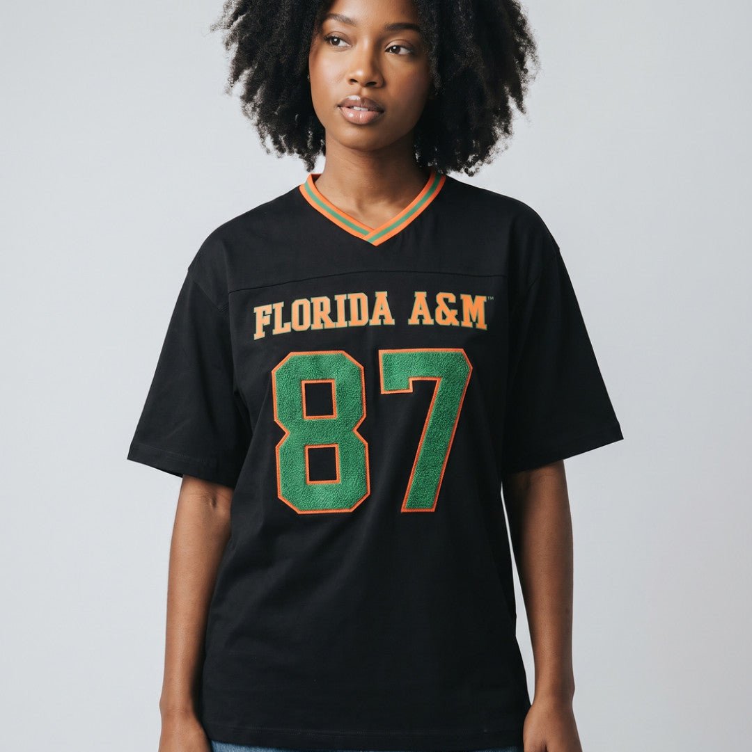 Florida A&M University Rattlers (FAMU) Oversized Jersey Tee (Lifestyle) - A woman wearing a black Florida A&M 87 jersey t-shirt with green and gold lettering, paired with blue jeans, against a light gray studio background.