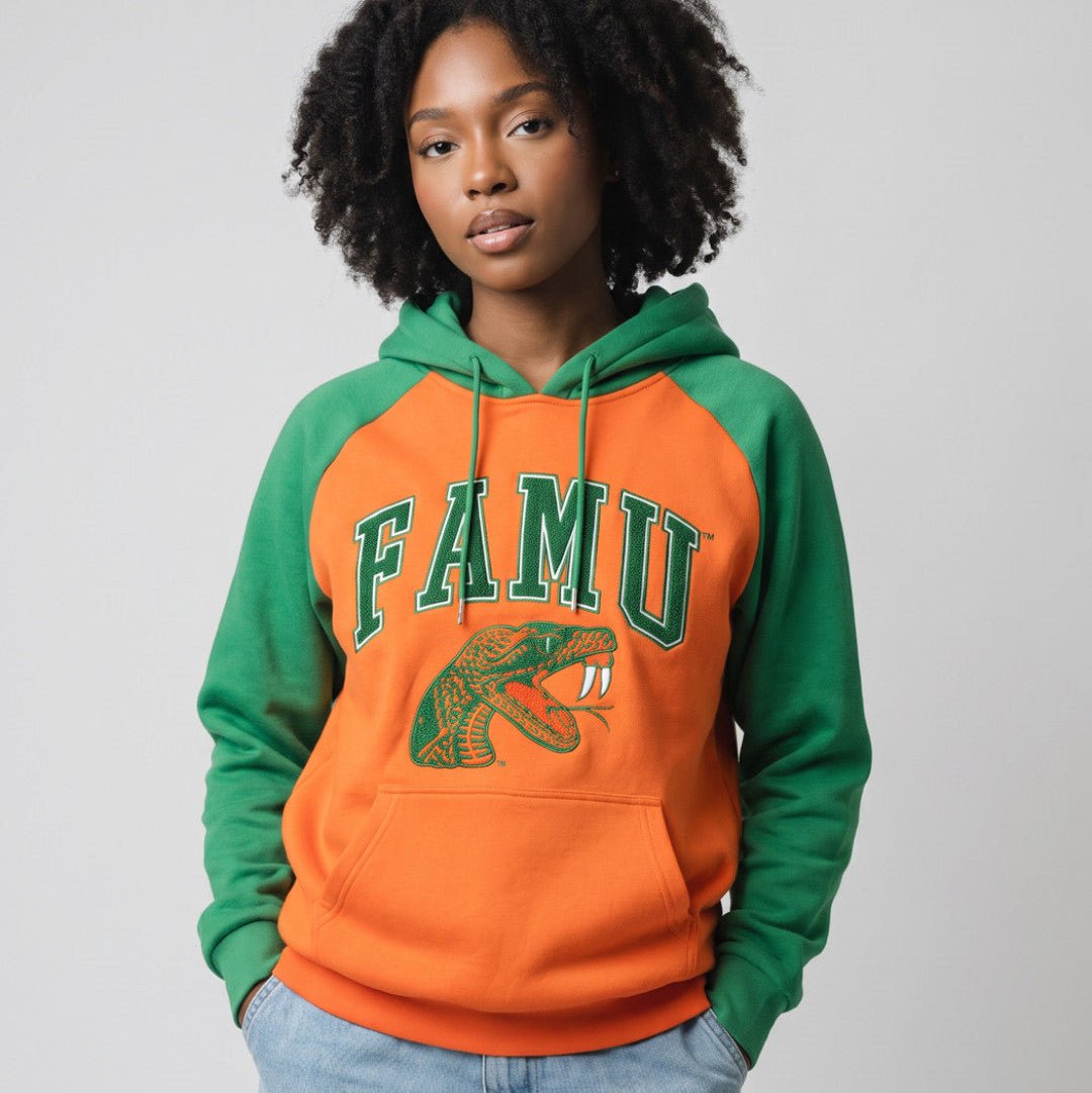 Florida A&M University (FAMU) Rattlers Hooded Sweatshirt (Lifestyle) - A woman wearing a two-tone orange and green raglan hoodie with 'FAMU' and a snake graphic on the front, paired with light blue jeans, against a plain gray studio background.