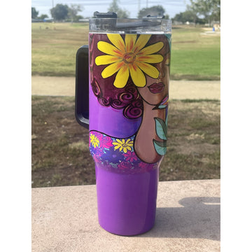 Garden Spirity by Pamela Hills: African American Tumbler with Long Handle (Llfestyle) - A purple insulated tumbler with a black handle and lid, decorated with a colorful floral and abstract illustration, photographed outdoors on a stone surface with a grassy field in the background.