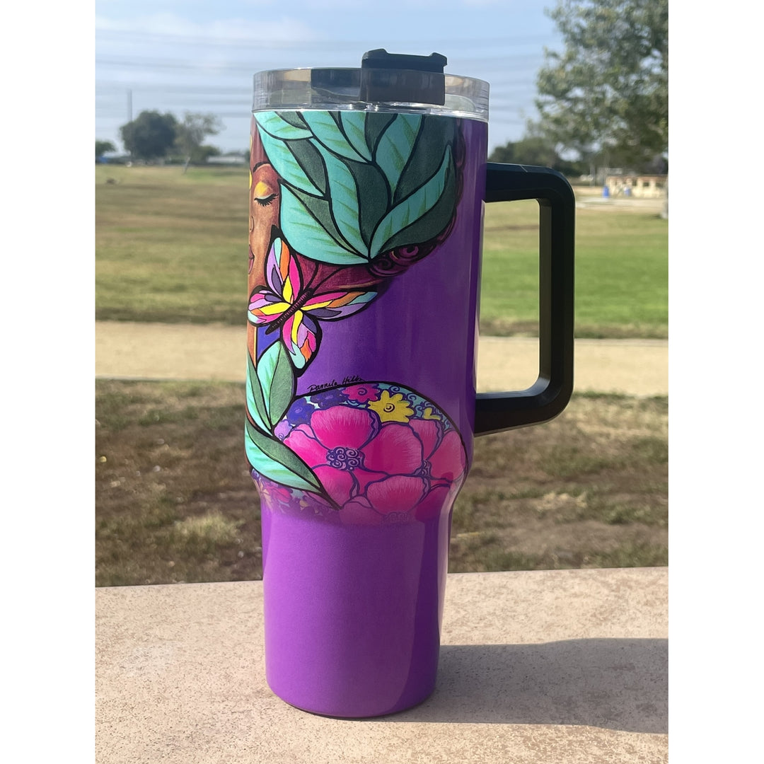 Garden Spirity by Pamela Hills: African American Tumbler with Long Handle (Llfestyle) - A purple travel tumbler with a black handle and lid, decorated with colorful floral and leaf patterns, photographed outdoors on a stone surface with a blurred park background.