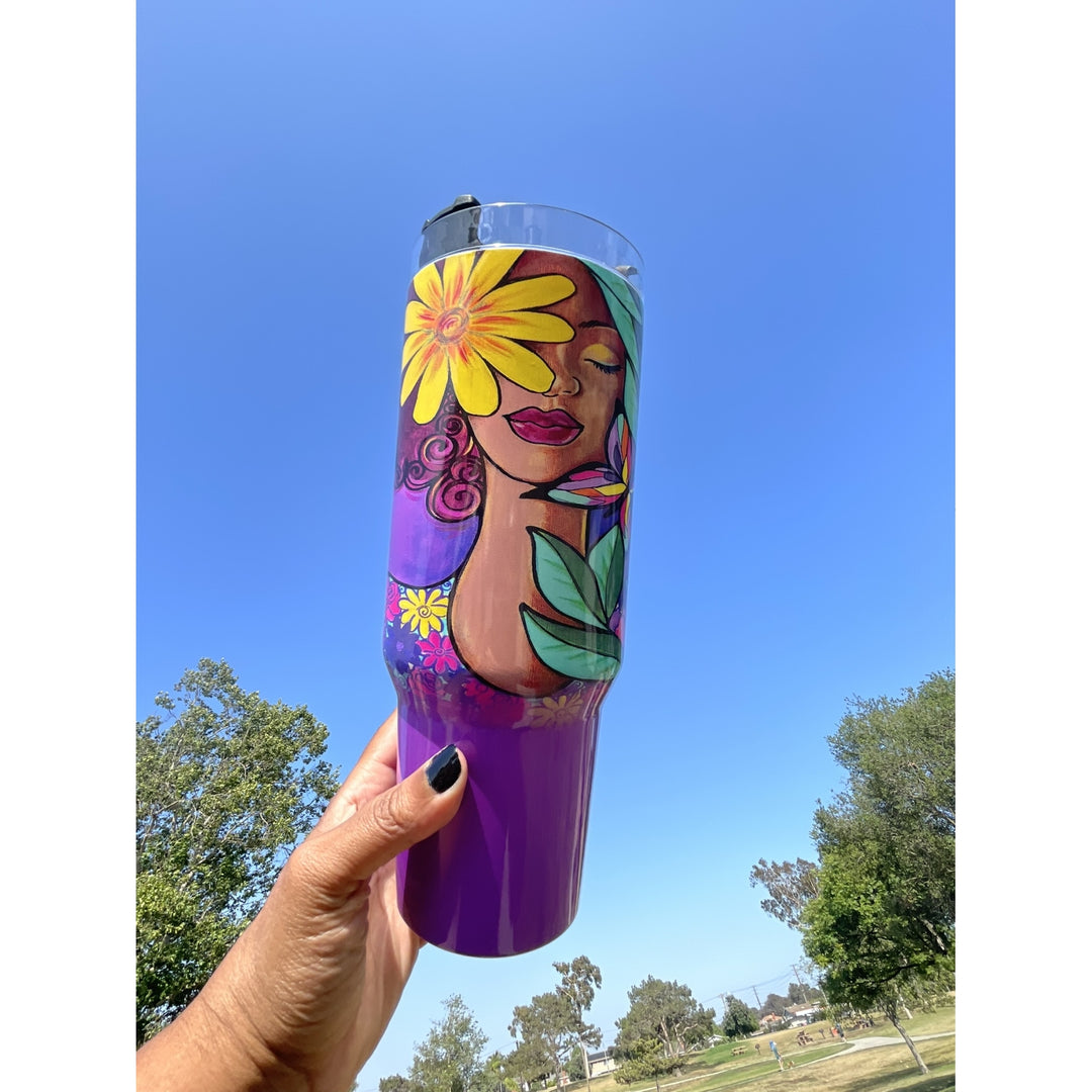 Garden Spirity by Pamela Hills: African American Tumbler with Long Handle (Llfestyle) - A hand with black nail polish holds a purple insulated tumbler featuring a colorful illustration of a woman with a yellow flower on her face, set against a blue sky and park background.