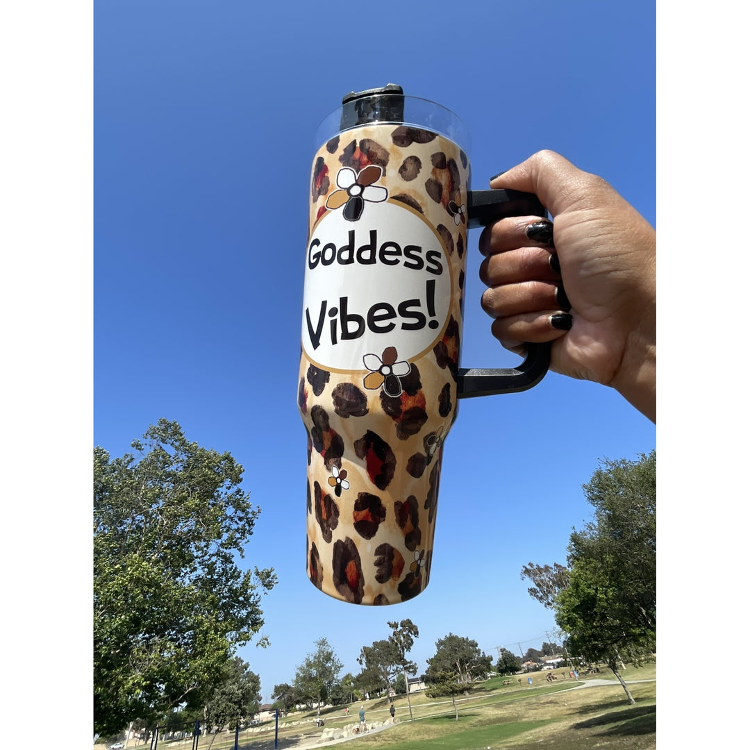 Goddess Vibes by Kiwi McDowell: African American Tumbler with Long Handle (Lifestyle) - A hand holding a leopard print tumbler with a black handle and lid, featuring a white oval label that reads 'Goddess Vibes!' with bee illustrations, set against an outdoor park background with blue sky.