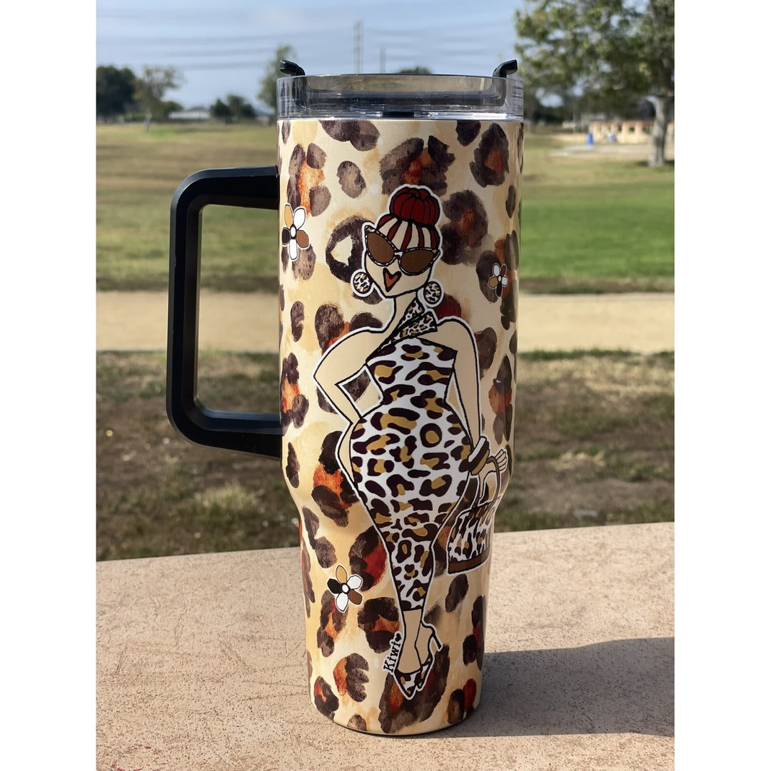 Goddess Vibes by Kiwi McDowell: African American Tumbler with Long Handle (Lifestyle) - A handled insulated tumbler with a beige leopard print pattern and a stylized woman in a leopard-print dress, photographed outdoors on a table with a park background.