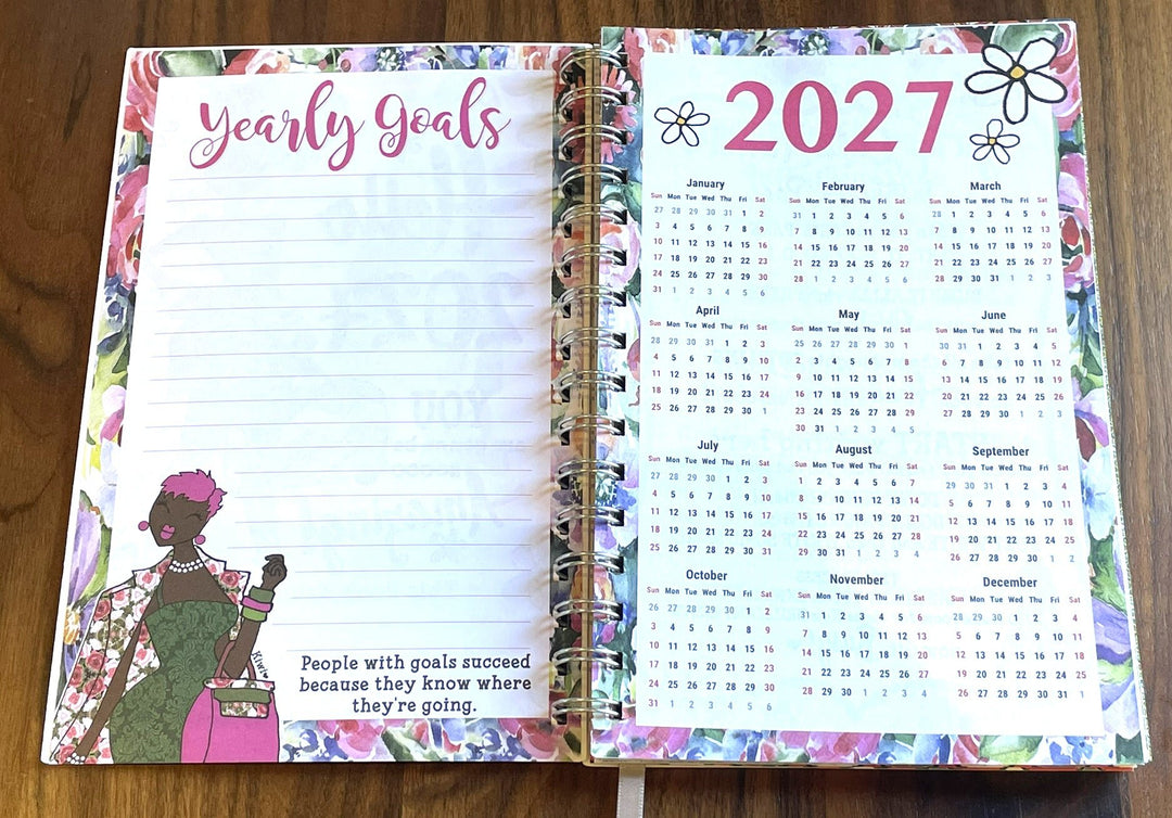 Good Vibes & Voice Mails 2027 Weekly Inspirational Planner by Kiwi McDowell (Yearly Goals) - An open spiral-bound planner on a wooden surface, showing the left page with lined writing space and the phrase 'Yearly Goals' in pink script, and the right page with a 2027 calendar layout. The planner has a colorful floral border and decorative illustrations.