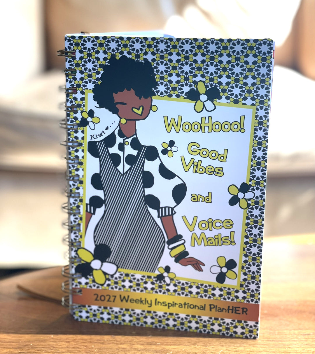 Good Vibes & Voice Mails 2027 Weekly Inspirational Planner by Kiwi McDowell (Lifestyle) - A spiral-bound 2027 Weekly Inspirational Planner titled 'Woohoo! Good Vibes and Voice Mail!' featuring a colorful cartoon illustration of a woman with an afro on a patterned cover, displayed on a wooden surface.