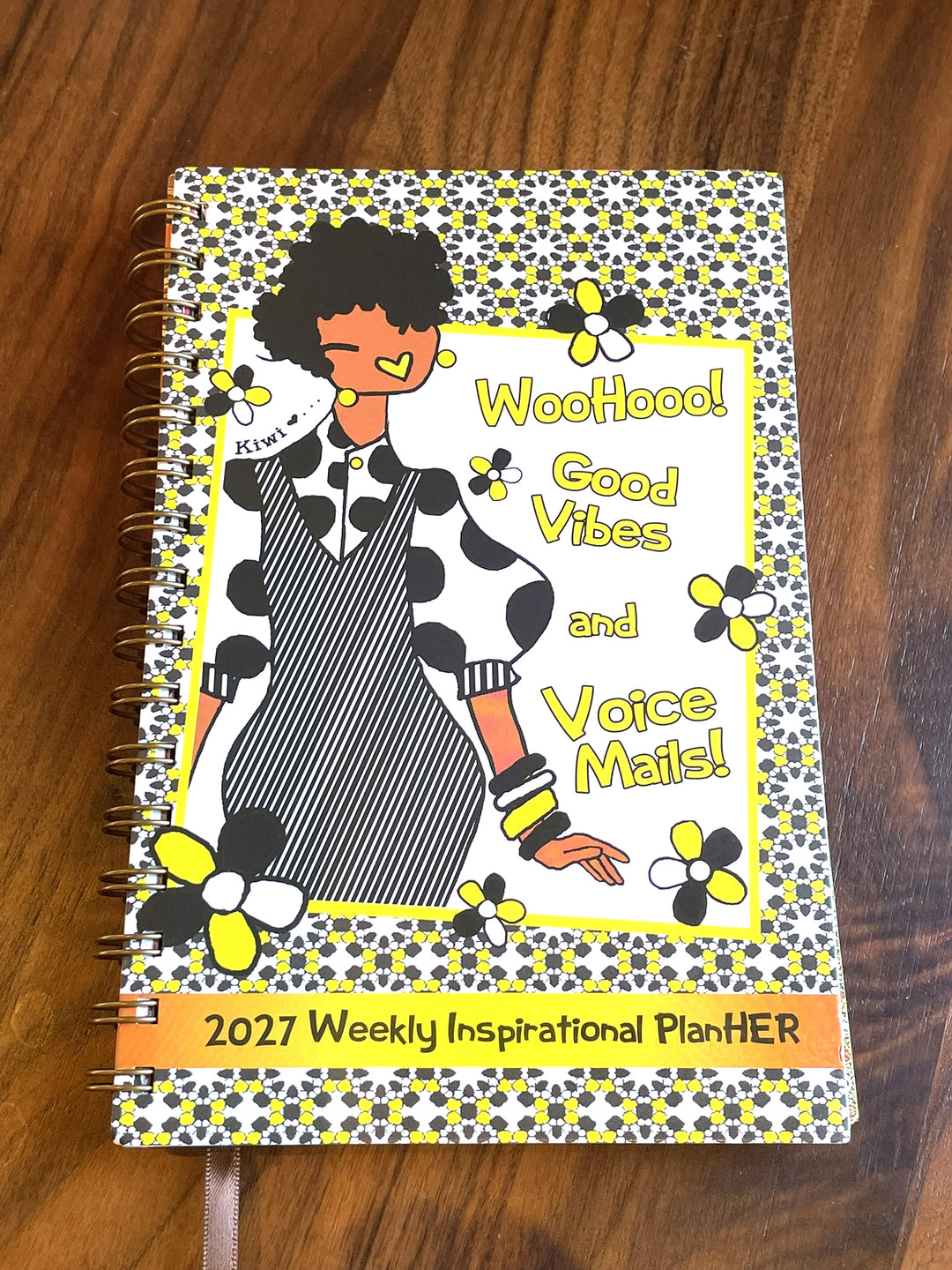 Good Vibes & Voice Mails 2027 Weekly Inspirational Planner by Kiwi McDowell (Lifestyle) - A 2027 Weekly Inspirational Planther notebook with a colorful cover illustration of a woman in a cow-print dress, placed on a wooden surface.