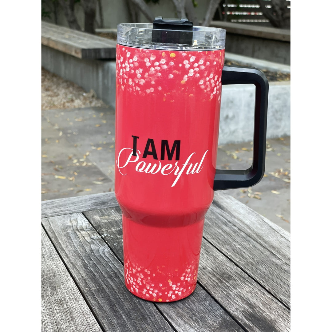 I am Powerful by Sylvia "GBaby" Cohen: Detla Sigma Theta Tumbler with Handle (Lifestyle) - A red insulated tumbler with a black handle and lid, featuring the text 'I AM Powerful' printed on the front, sitting on a weathered wooden table outdoors.