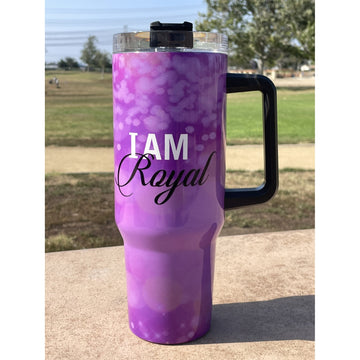 I am Royal by Sylvia "GBaby" Cohen: African American 40 Ounce Tumbler with Long Handle (Lifestyle) - A purple and lavender gradient travel tumbler with a black handle and lid, featuring the text 'I AM Royal' printed on the front, set outdoors on a concrete surface with a park in the background.