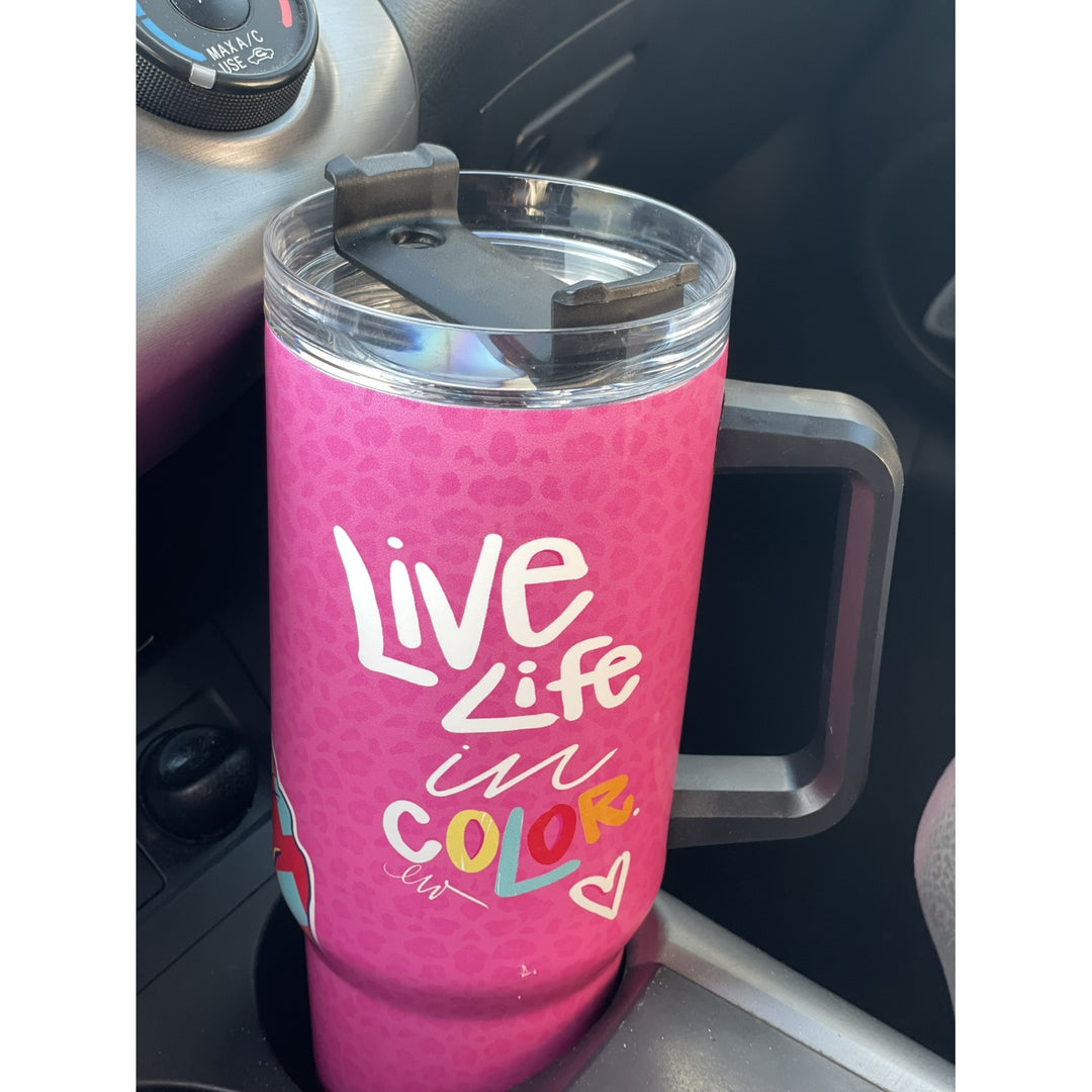 Live Life in Color by Madeleine Wood: African American Tumbler with Handle (Lifestyle) - A pink handled travel tumbler with a clear lid sits in a car cup holder. The tumbler has a subtle leopard print texture and is printed with the phrase 'Live Life in COLOR' in white, yellow, and blue lettering.