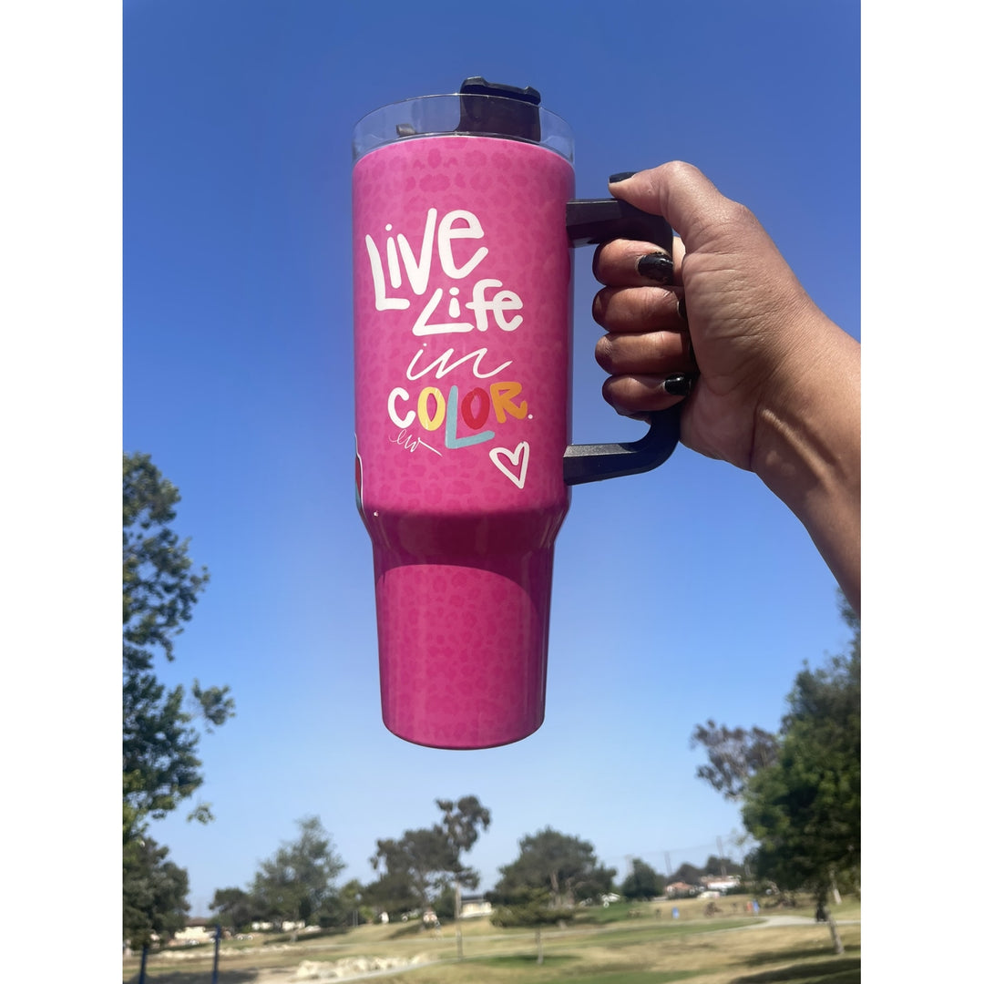 Live Life in Color by Madeleine Wood: African American Tumbler with Handle (Lifestyle) - A hand holds a bright pink insulated tumbler with a black handle and lid against a blue sky and park background. The tumbler has white and multicolor text that reads 'Live Life in COLOR' with a small heart.