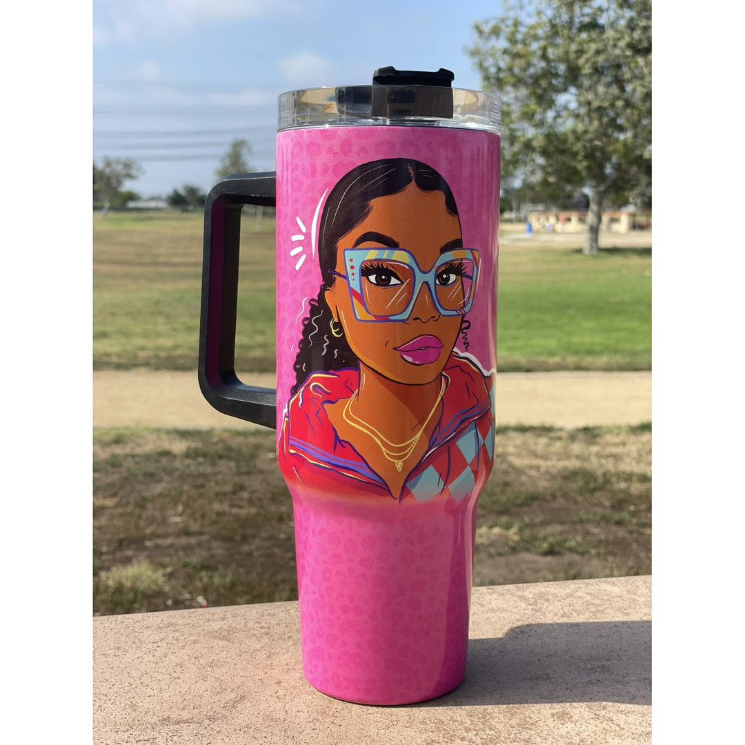 Live Life in Color by Madeleine Wood: African American Tumbler with Handle (Lifestyle) - A tall pink insulated travel tumbler with a black handle and lid, featuring a colorful illustrated portrait of a woman with glasses on the front, photographed outdoors on a stone surface with a blurred park background.