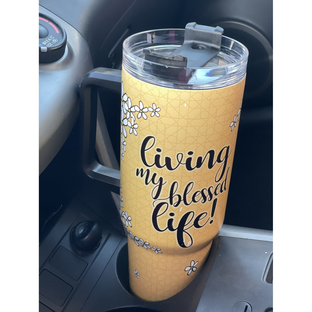 Living My Blessed Life by Kiwi McDowell: African American Tumbler with Long Handle (Lifestyle) - A yellow insulated travel tumbler with a clear lid and black handle, featuring the text 'living my blessed life!' in black script and small white daisy illustrations, shown sitting in a car cup holder.
