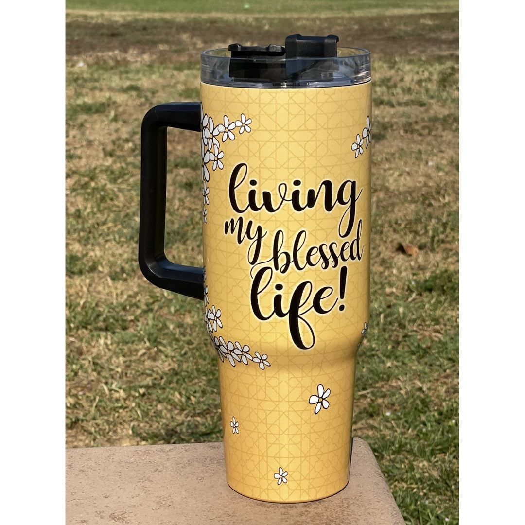 Living My Blessed Life by Kiwi McDowell: African American Tumbler with Long Handle (Lifestyle) - A yellow insulated travel tumbler with a black handle and lid, featuring the phrase 'living my blessed life!' in black script and small white floral illustrations, photographed outdoors on a stone surface with grass in the background.