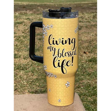 Living My Blessed Life by Kiwi McDowell: African American Tumbler with Long Handle (Lifestyle) - A yellow insulated travel tumbler with a black handle and lid, featuring the phrase 'living my blessed life!' in black script and small white floral illustrations, photographed outdoors on a stone surface with grass in the background.