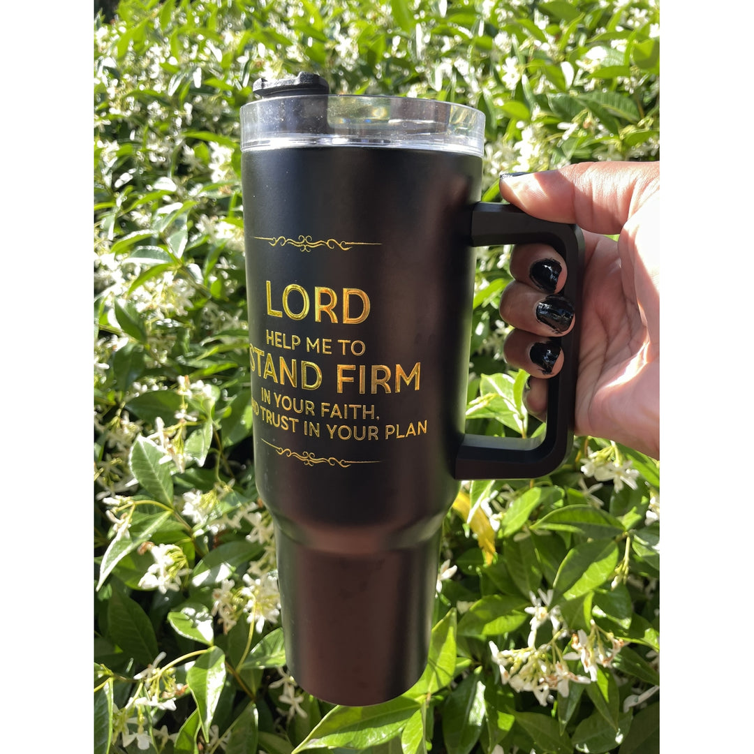 Lord Help Me Stand Firm: African American Faith Tumbler with Handle (Lifestyle) - A black insulated travel tumbler with a clear lid and black handle, held by a hand against a background of green foliage. The tumbler features gold printed text that reads: 'LORD HELP ME TO STAND FIRM IN YOUR FAITH TRUST IN YOUR PLAN'.