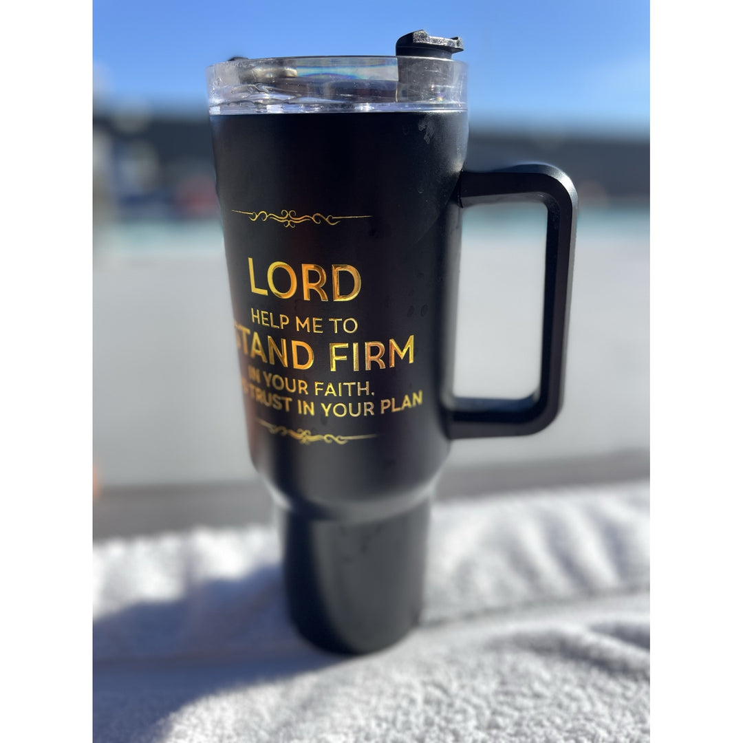 Lord Help Me Stand Firm: African American Faith Tumbler with Handle (Lifestyle) - A black insulated travel tumbler with a clear lid and black handle, featuring gold text that reads 'LORD HELP ME TO STAND FIRM' and 'TRUST IN YOUR PLAN,' shown outdoors on a towel with a blurred background.