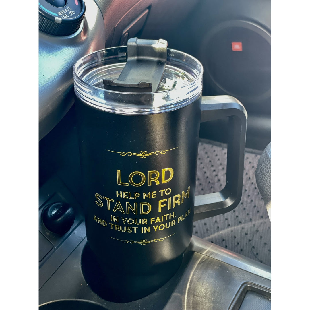 Lord Help Me Stand Firm: African American Faith Tumbler with Handle (Lifestyle) - A black insulated travel mug with a handle and clear lid, placed in a car cup holder. The mug has gold text that reads, 'LORD HELP ME TO STAND FIRM IN YOUR FAITH AND TRUST IN YOUR PLACES.