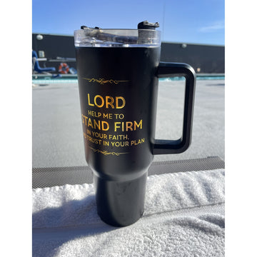 Lord Help Me Stand Firm: African American Faith Tumbler with Handle (Lifestyle) - A black insulated travel tumbler with a clear lid and black handle, standing on a white towel outdoors. The tumbler has gold text that reads, 'LORD HELP ME TO STAND FIRM IN YOUR FAITH AND TRUST IN YOUR PLAN.