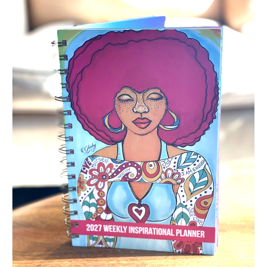 Love Yourself 2027 Weekly Inspirational Planner by Sylvia "Gbaby" Cohen (Lifestyle) - A spiral-bound 2027 Weekly Inspirational Planner notebook featuring a colorful illustration of a woman with a large pink afro on the cover, standing on a wooden surface.