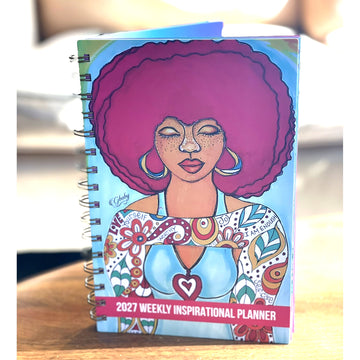 Love Yourself 2027 Weekly Inspirational Planner by Sylvia "Gbaby" Cohen (Lifestyle) - A spiral-bound 2027 Weekly Inspirational Planner notebook featuring a colorful illustration of a woman with a large pink afro on the cover, standing on a wooden surface.