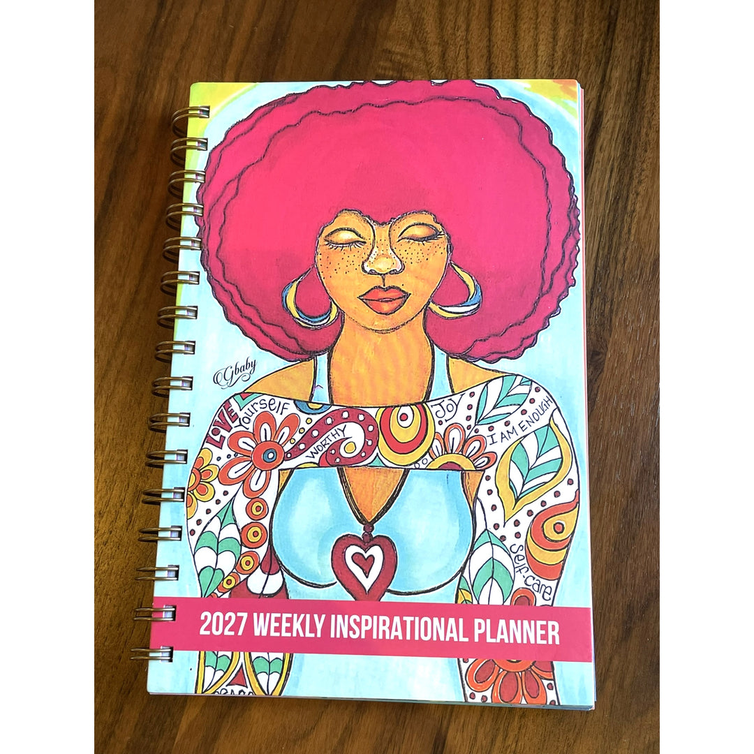 Love Yourself 2027 Weekly Inspirational Planner by Sylvia "Gbaby" Cohen (Lifestyle 2) - A spiral-bound 2027 Weekly Inspirational Planner notebook featuring a colorful illustration of a woman with a pink afro on a wooden table background.