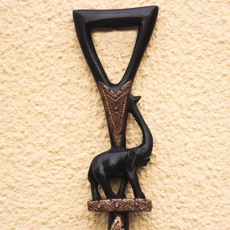 Loyalty and Community (Strolling Elephant): Decorative African Wooden Walking Stick (Main, Detail) - A dark carved wooden wall decor piece featuring an elephant and a decorative triangular top, mounted against a textured beige stucco wall.