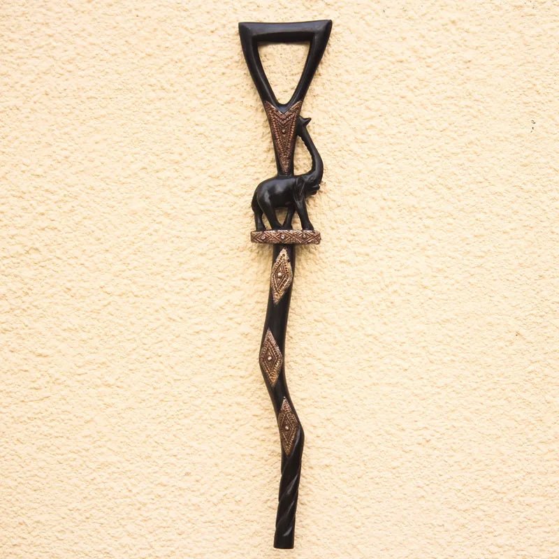 Loyalty and Community (Strolling Elephant): Decorative African Wooden Walking Stick (Main) - A dark carved wall decor piece featuring a stylized elephant on a staff with decorative bronze-toned geometric accents, mounted against a textured beige wall.
