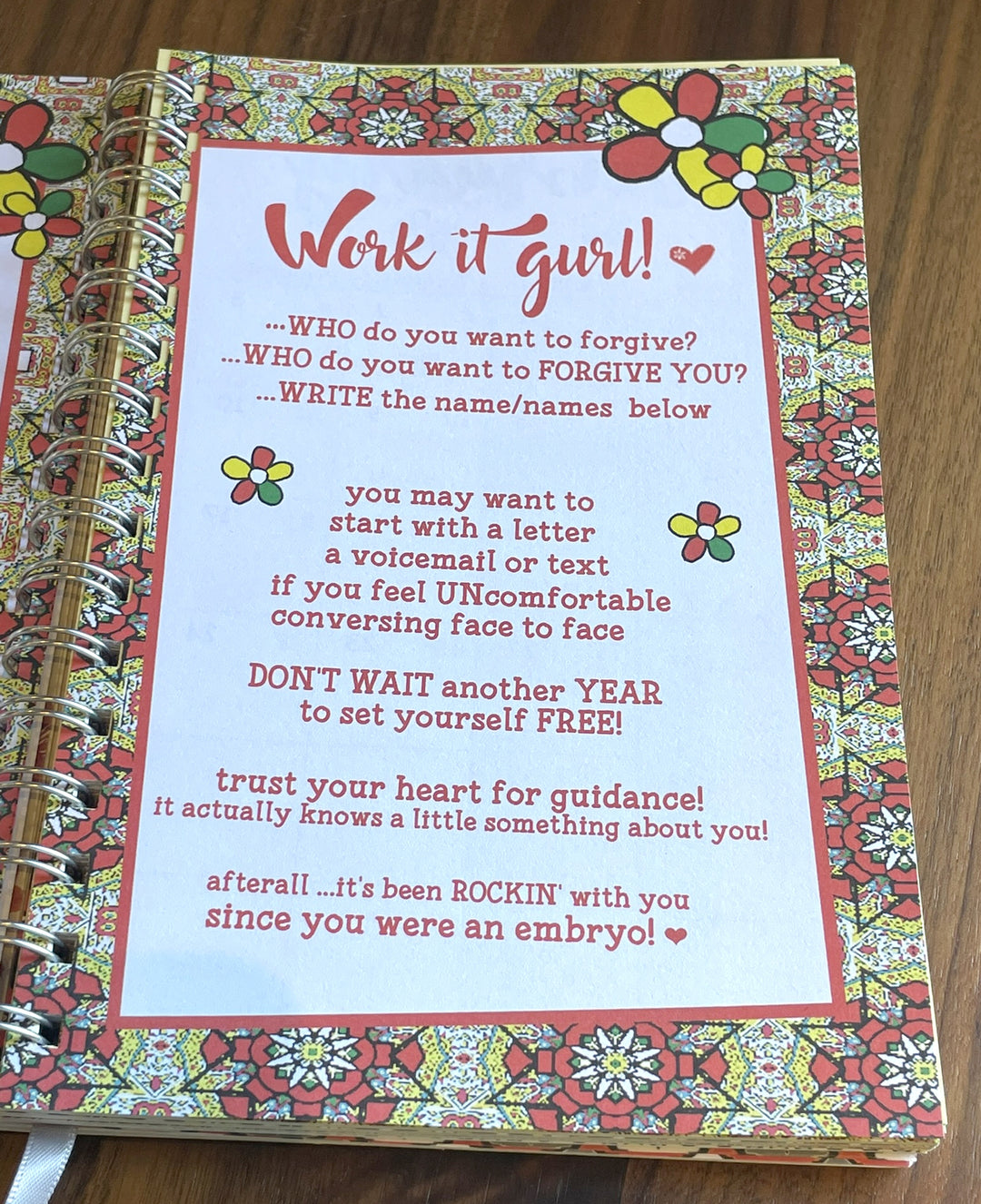 Net Worth and Nature Trails 2027 Weekly Inspirational Planner by Kiwi McDowell (Affirmation Pages III) - A spiral-bound journal or notebook with a colorful floral patterned cover and a large white text insert that reads 'Work it gurl!' with motivational self-improvement text on a wooden surface.