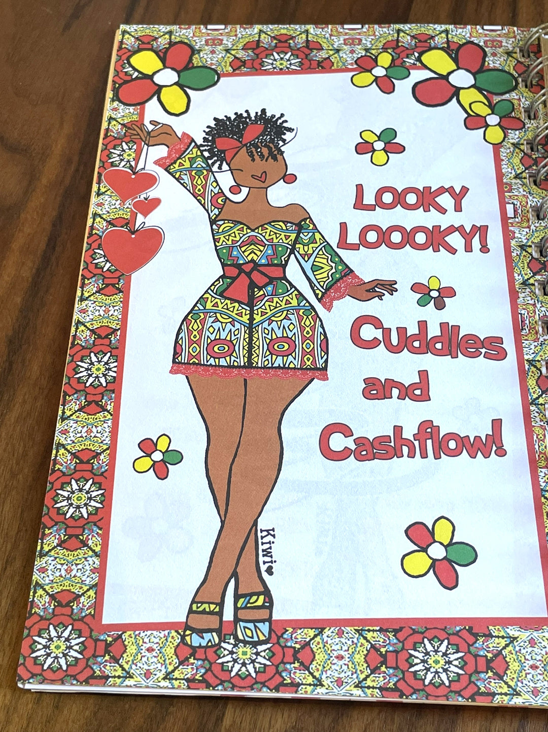 Net Worth and Nature Trails 2027 Weekly Inspirational Planner by Kiwi McDowell (Art Pages) - A square printed card or cover featuring a colorful illustrated woman in a patterned dress with the text 'Looky looney! Cuddles and Cashflow!' on a white center panel, surrounded by a red and multicolor floral border, photographed on a wooden surface.