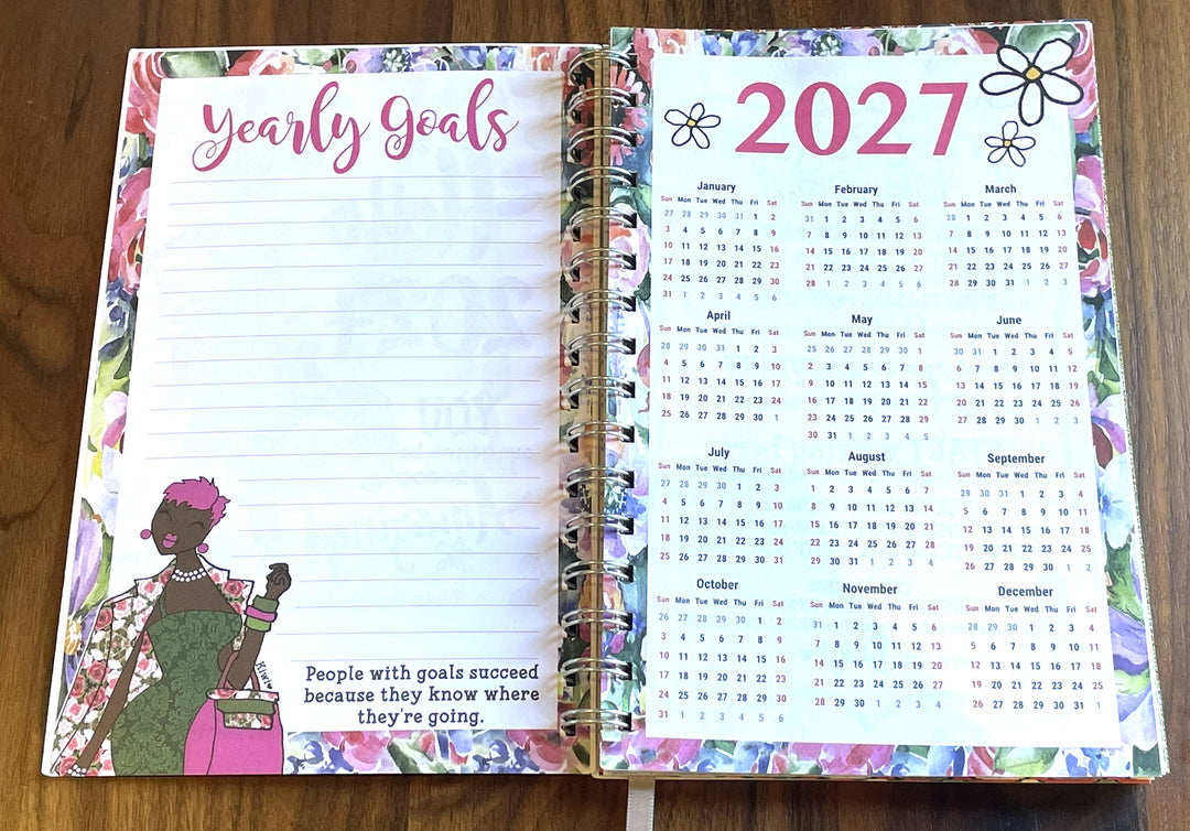 Net Worth and Nature Trails 2027 Weekly Inspirational Planner by Kiwi McDowell (Yearly Goals) - An open spiral-bound planner on a wooden surface, showing the left page with lined writing space and the phrase 'Yearly Goals' in pink script, and the right page with a 2027 calendar layout. The planner has a colorful floral border and decorative illustrations.
