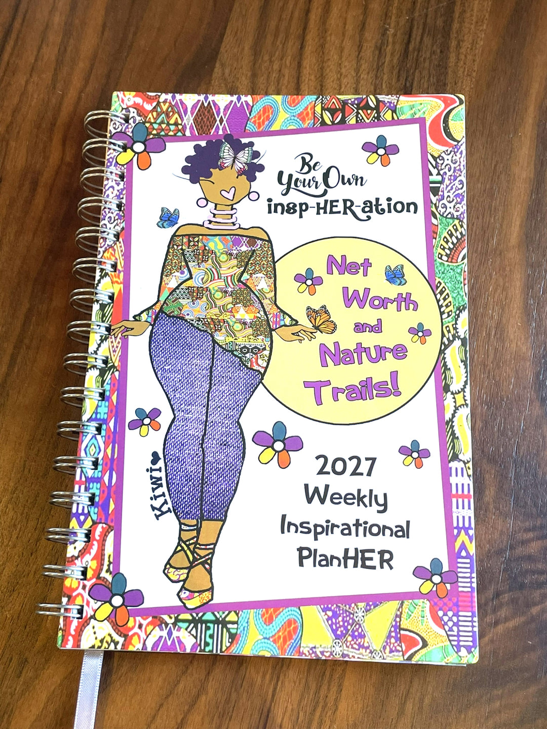 Net Worth and Nature Trails 2027 Weekly Inspirational Planner by Kiwi McDowell (Lifestyle II) - A spiral-bound 2027 weekly inspirational planner titled 'Be Your Own inspiration. Not Worth and Nature Trails!' lies open on a dark wooden table.