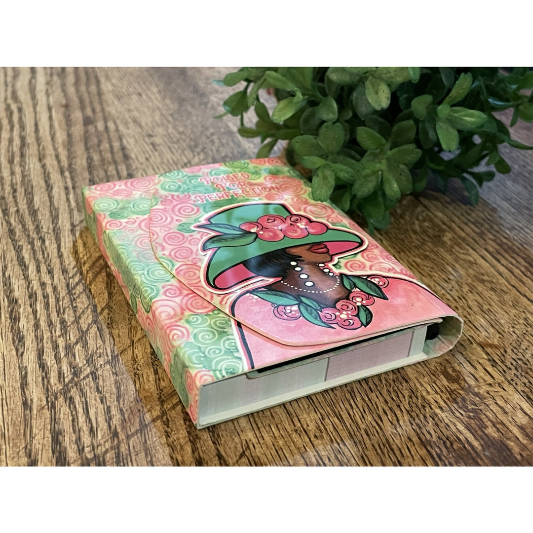 Poised for Perfection (Alpha Kappa Alpha) Stylus Purse Pal by Pamela Hills (Lifestyle 2) - Decorative notebook with a colorful design on a wooden surface with a plant in the background