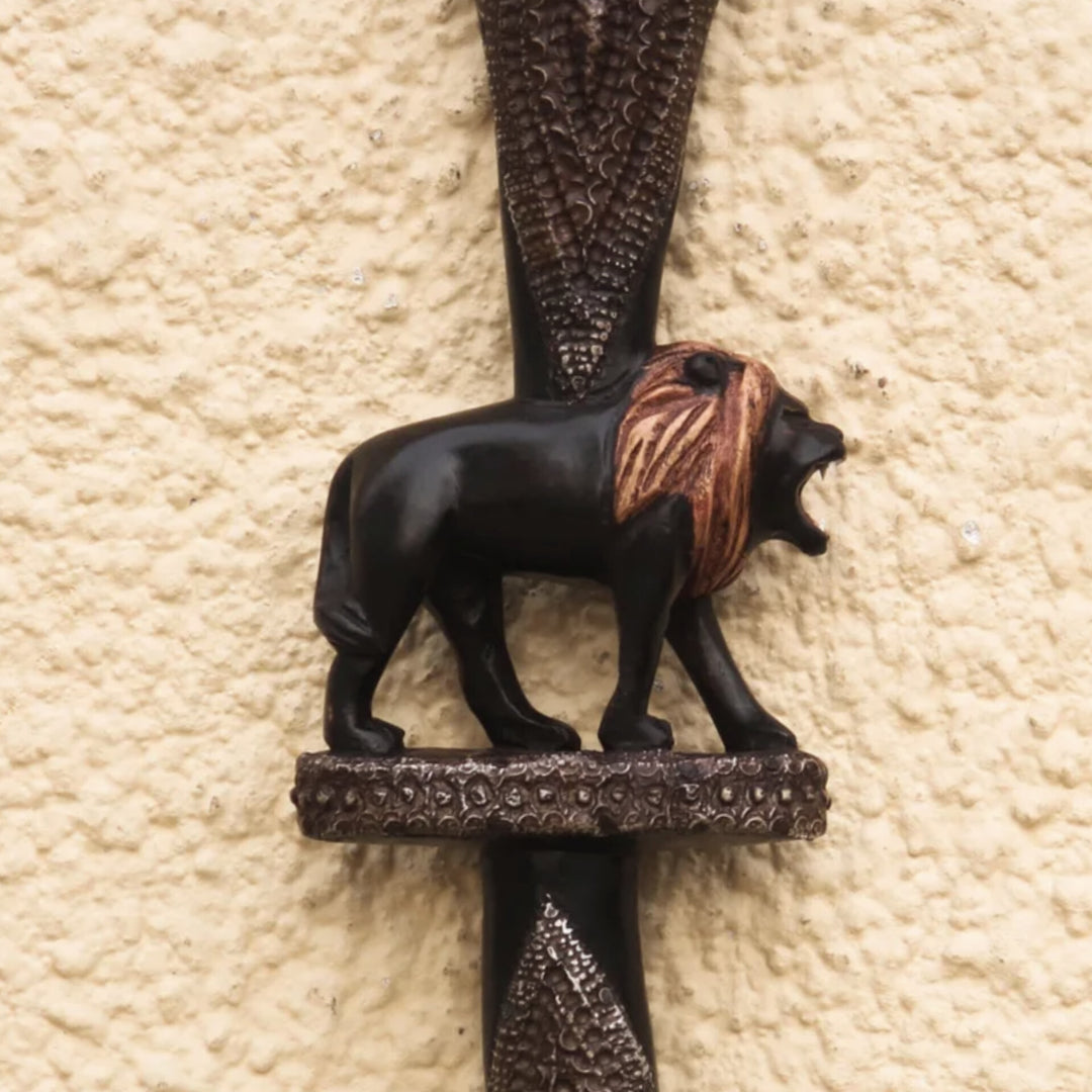 Royalty and Courage (Strolling Lion): Decorative African Wooden Walking Stick (Main, Detail) - A decorative lion wall plaque mounted on a textured beige stucco wall. The lion figurine is black with a brown and tan mane, standing on an ornate dark metal crossbar.