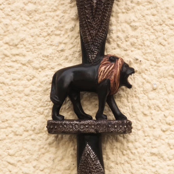 Royalty and Courage (Strolling Lion): Decorative African Wooden Walking Stick (Main, Detail) - A decorative lion wall plaque mounted on a textured beige stucco wall. The lion figurine is black with a brown and tan mane, standing on an ornate dark metal crossbar.