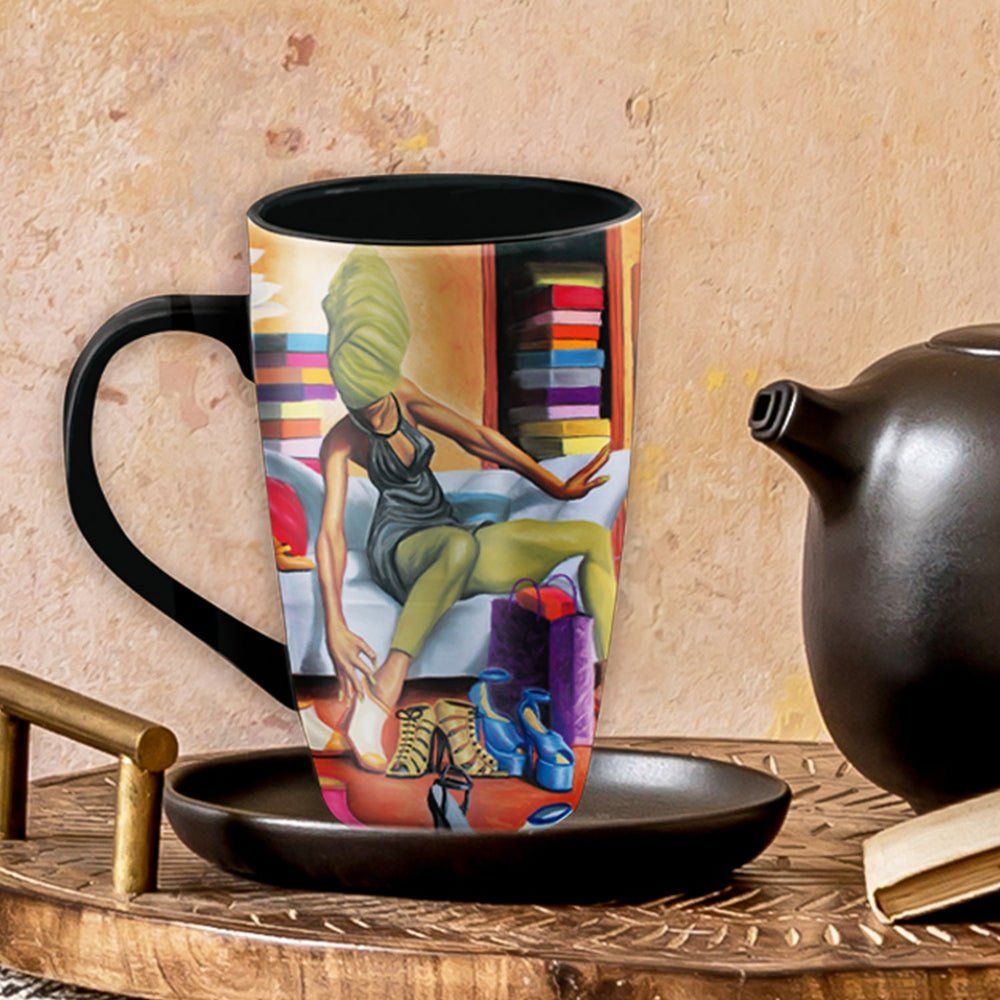 Shoe Addict Ceramic Latte Mug by Dion Pollard (Lifestyle) - A tall ceramic mug with a black handle and interior sits on a matching black saucer on a wooden table. The mug is printed with a colorful artistic illustration of a woman in green and red clothing.