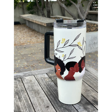 Sister Strong by Janine Robinson: African American 40 Ounce Tumbler with Handle (Lifestyle) - A white insulated tumbler with a black handle and lid sits on a weathered wooden table outdoors. The tumbler features a printed design of three Black women with red clothing under gray leaves and small yellow flowers.