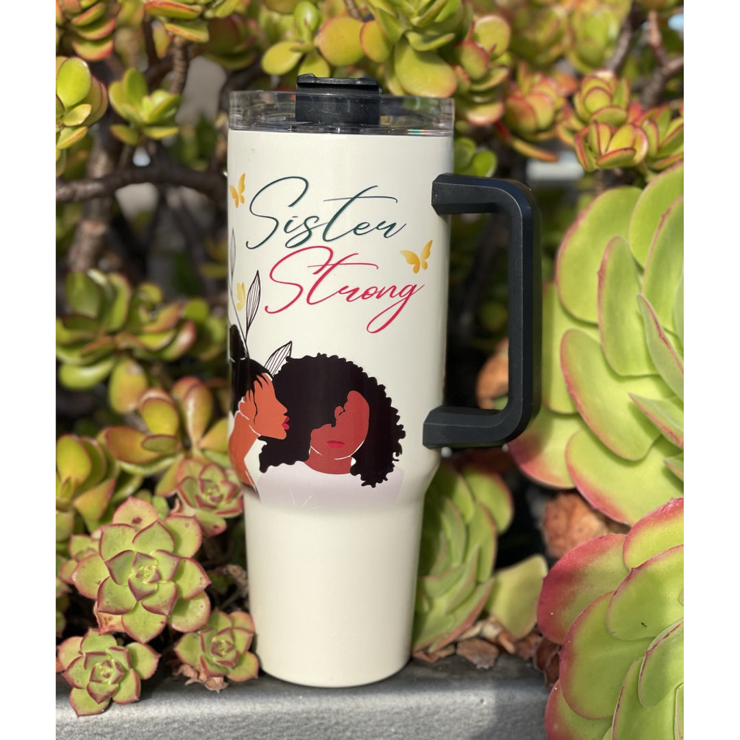 Sister Strong by Janine Robinson: African American 40 Ounce Tumbler with Handle (Lifestyle) - A white insulated travel tumbler with a black handle and lid, featuring the text 'Sister Strong' in teal and pink script with a graphic of two Black women embracing. The tumbler is photographed outdoors against a background of green succulents.