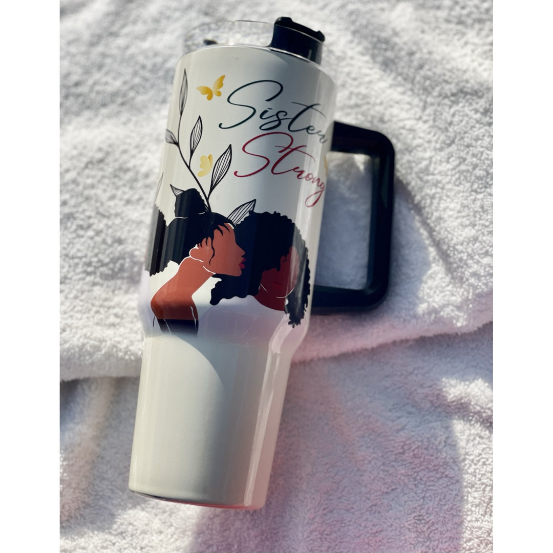 Sister Strong by Janine Robinson: African American 40 Ounce Tumbler with Handle (Lifestyle) - A white insulated tumbler with a black lid, featuring a printed design of two Black women embracing with the words 'Sister Strong' in black and red script, resting on a white towel with a black mirror behind it.