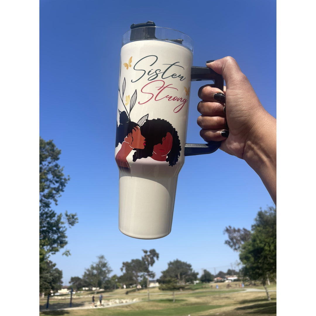 Sister Strong by Janine Robinson: African American 40 Ounce Tumbler with Handle (Lifestyle) - A hand holds a tall insulated tumbler outdoors against a blue sky and park background. The tumbler is white with a black handle and lid, decorated with the words 'Sister Strong' and an illustration of a Black woman with dark hair and red/orange accents.