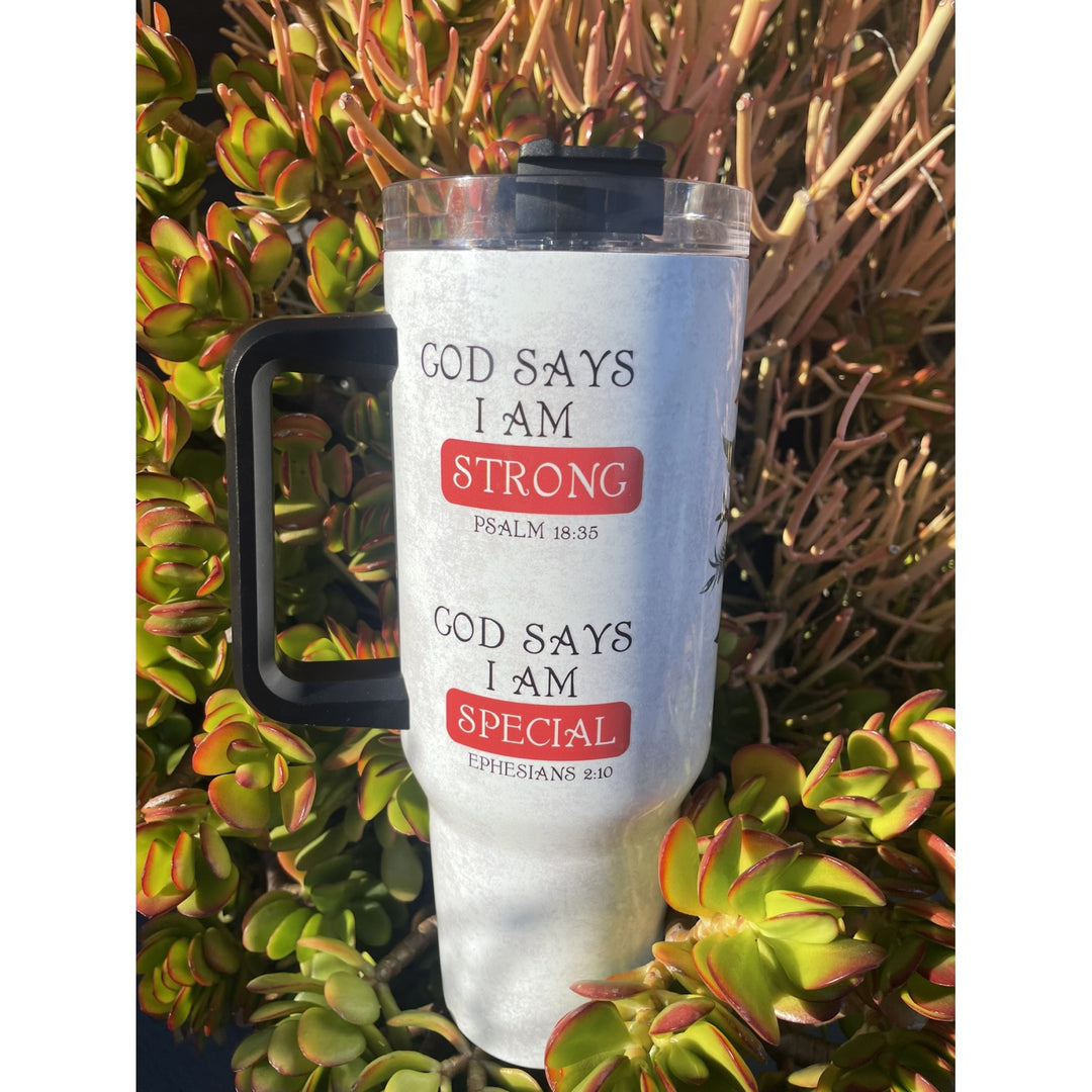 Strong and Special: African American Religious/Faith Tumbler with Handle (Lifestyle) - A white insulated tumbler with a black handle and lid, featuring the text 'GOD SAYS I AM STRONG' and 'GOD SAYS I AM SPECIAL' in black lettering with red rectangular highlights. The tumbler is photographed outdoors against a background of green and reddish succulent plants.