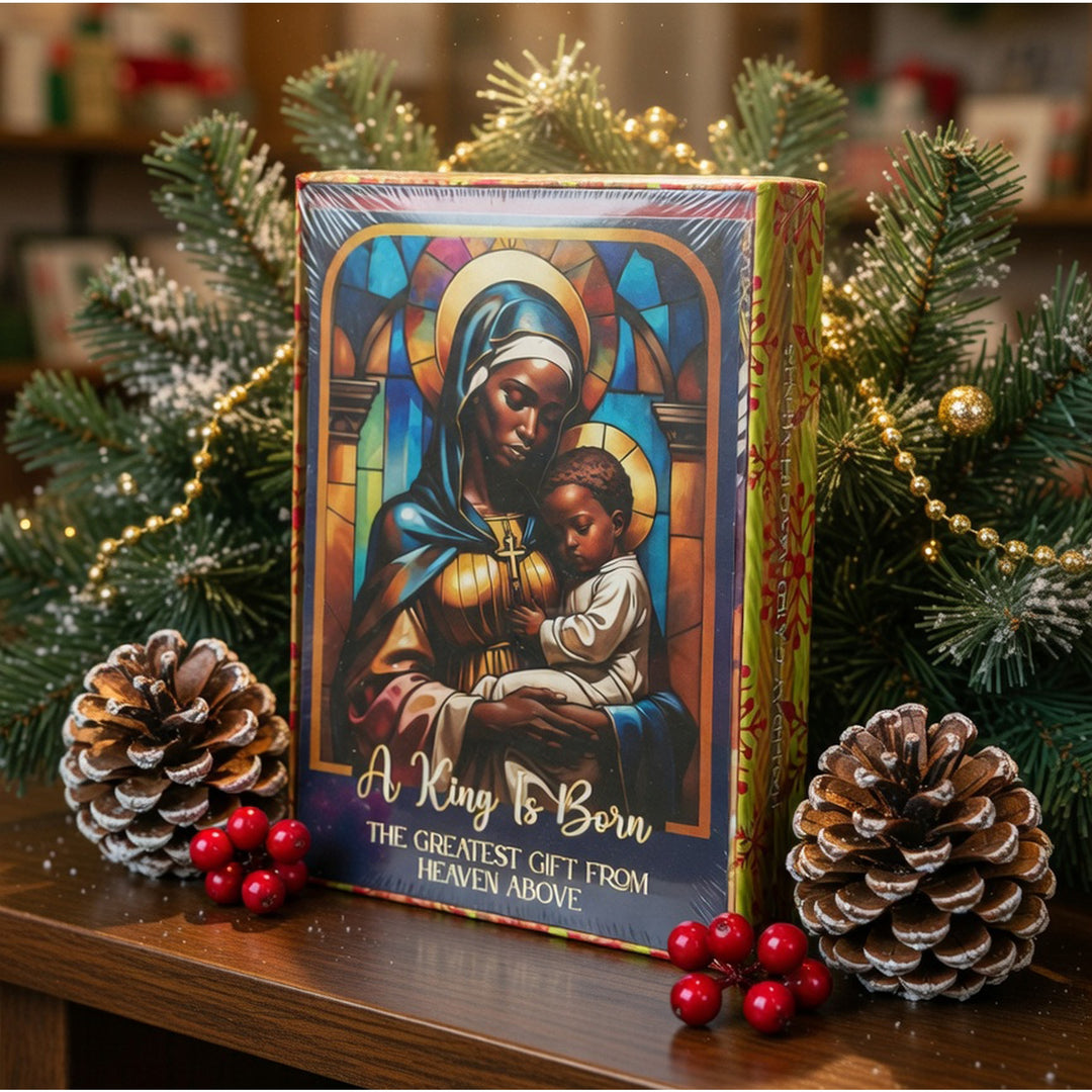 The Greatest Gift: African American Christmas Card Box Set by Janine Kai Robinson (Lifestyle) - A Christmas-themed greeting card or gift box featuring a stained-glass-style illustration of the Virgin Mary holding a baby Jesus, with the text 'A King Is Born' and 'The Greatest Gift From Heaven Above.' It is styled on a wooden surface with pine branches, pinecones, and red berries in a festive indoor setting.