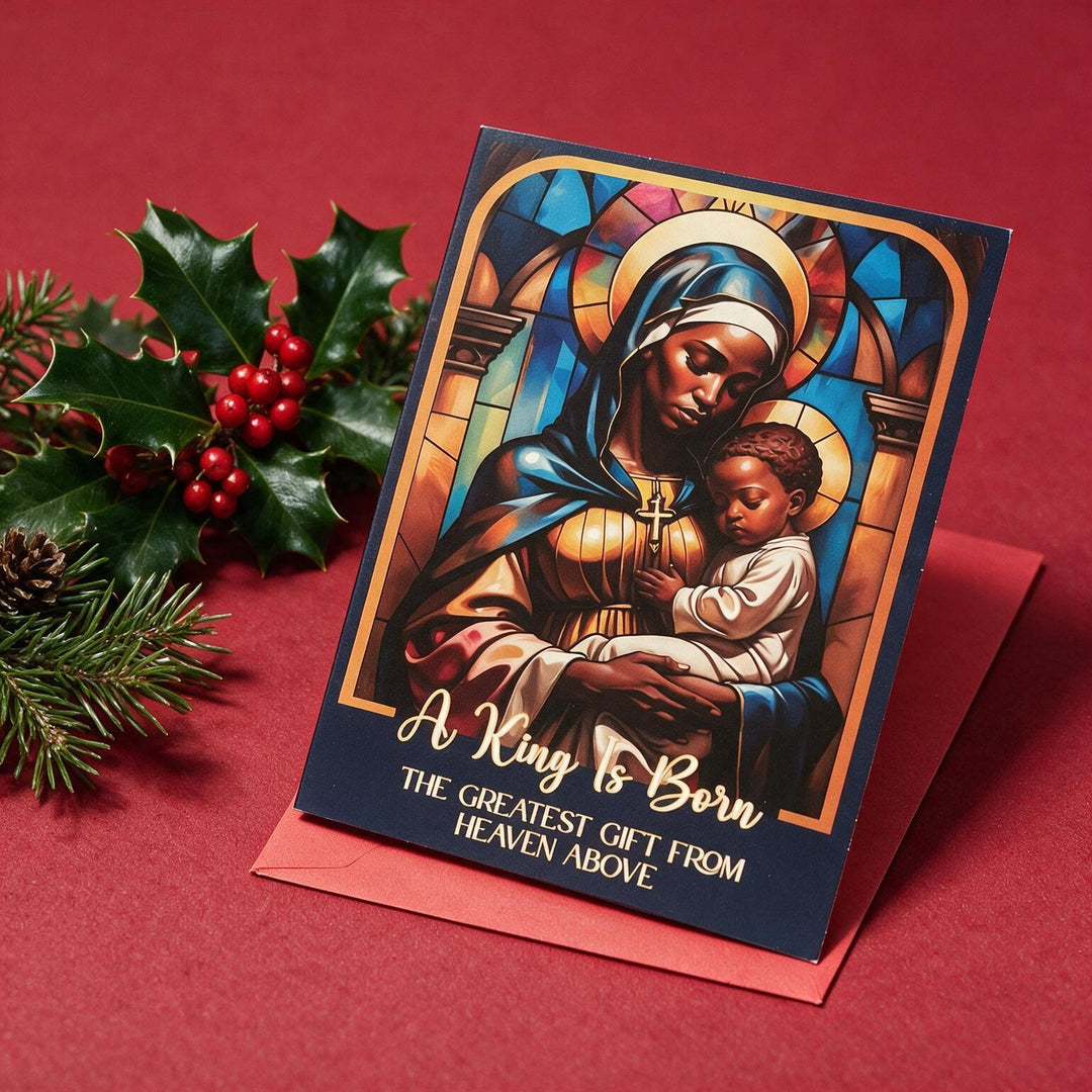 The Greatest Gift: African American Christmas Card Box Set by Janine Kai Robinson (Lifestyle 2) - A Christmas-themed greeting card featuring a stained-glass style illustration of Mary holding baby Jesus, with the text 'A King Is Born' and 'THE GREATEST GIFT FROM HEAVEN ABOVE,' displayed on a red background with holly and pine branches.