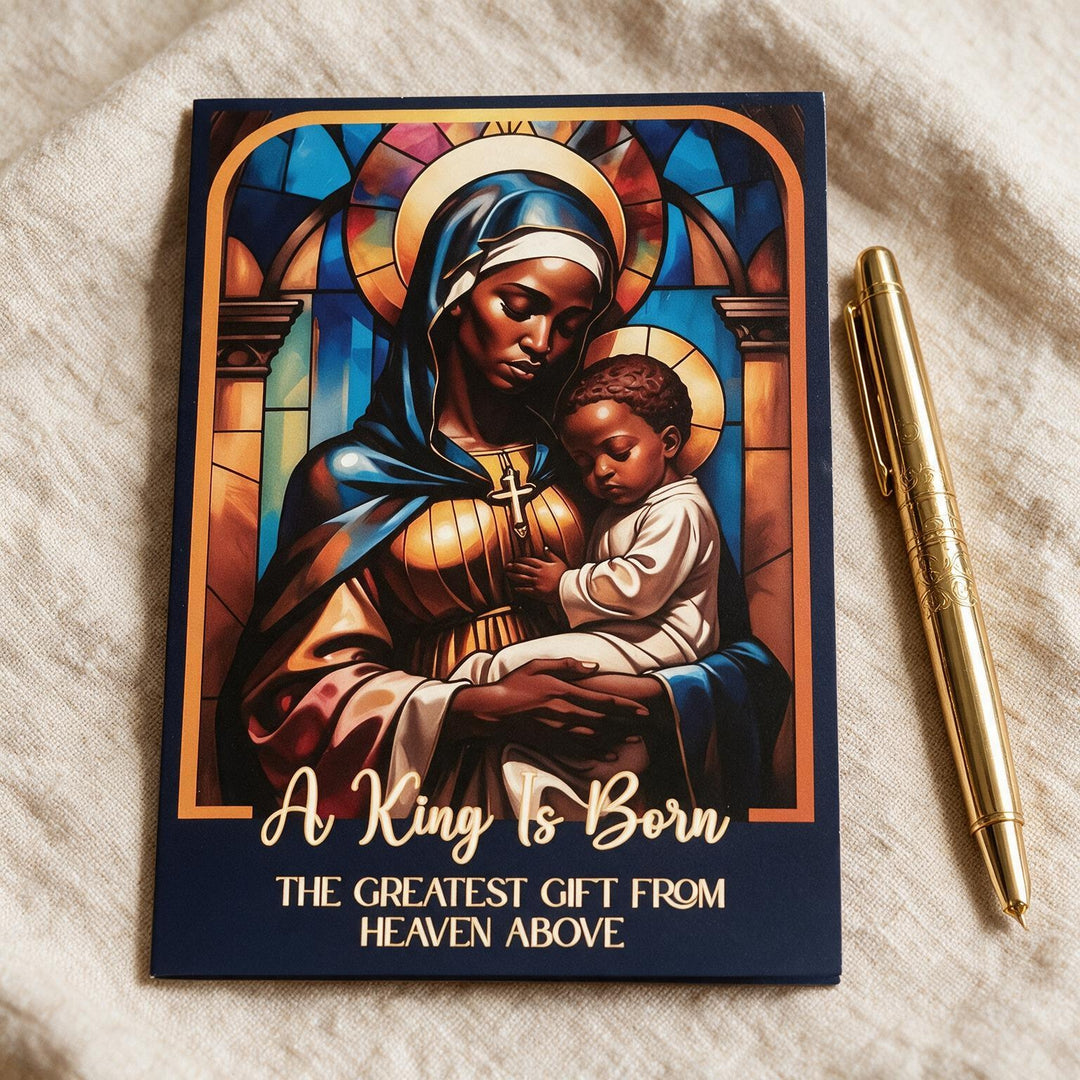 The Greatest Gift: African American Christmas Card Box Set by Janine Kai Robinson (Lifestyle 3) - A rectangular printed card or gift tag featuring a stained-glass-style illustration of a woman holding a child, with the text 'A King Is Born' and 'THE GREATEST GIFT FROM HEAVEN ABOVE,' placed on a beige fabric surface next to a gold pen.